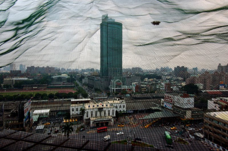 城市印象  張家豪<br>地點：台南火車站前大樓頂<br/>從大樓樓頂透過綠色護網俯看整個火車站，與後方的地標遠東百貨大樓，以這張作為整個系列的第一張相片，是個很棒的開始。<br/>